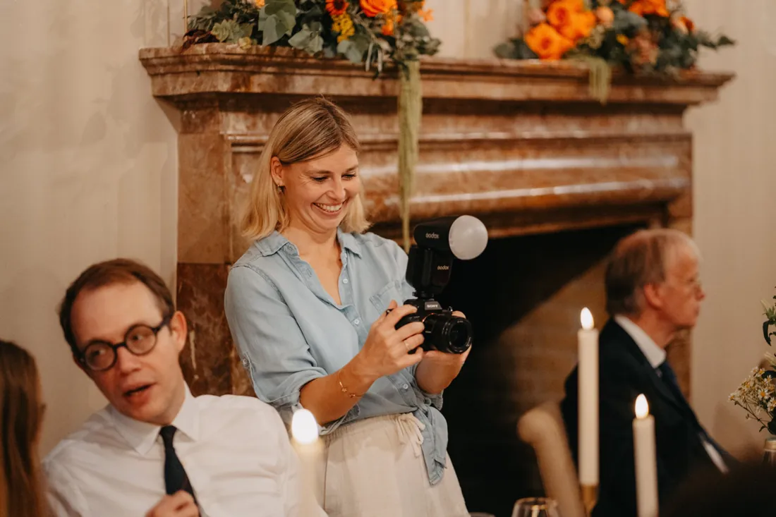 Lisa lacht mit Kamera und Blitz vor einem Kamin bei einer Hochzeitsfeier